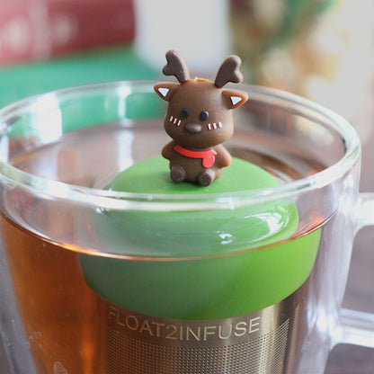 cute brown reindeer with a red scarf tea infuser for loose leaf tea with a fine mesh stainless steel tea strainer that reads FLOAT2INFUSE on a dark green base with books, a tea kettle, and christmas decorations in the background