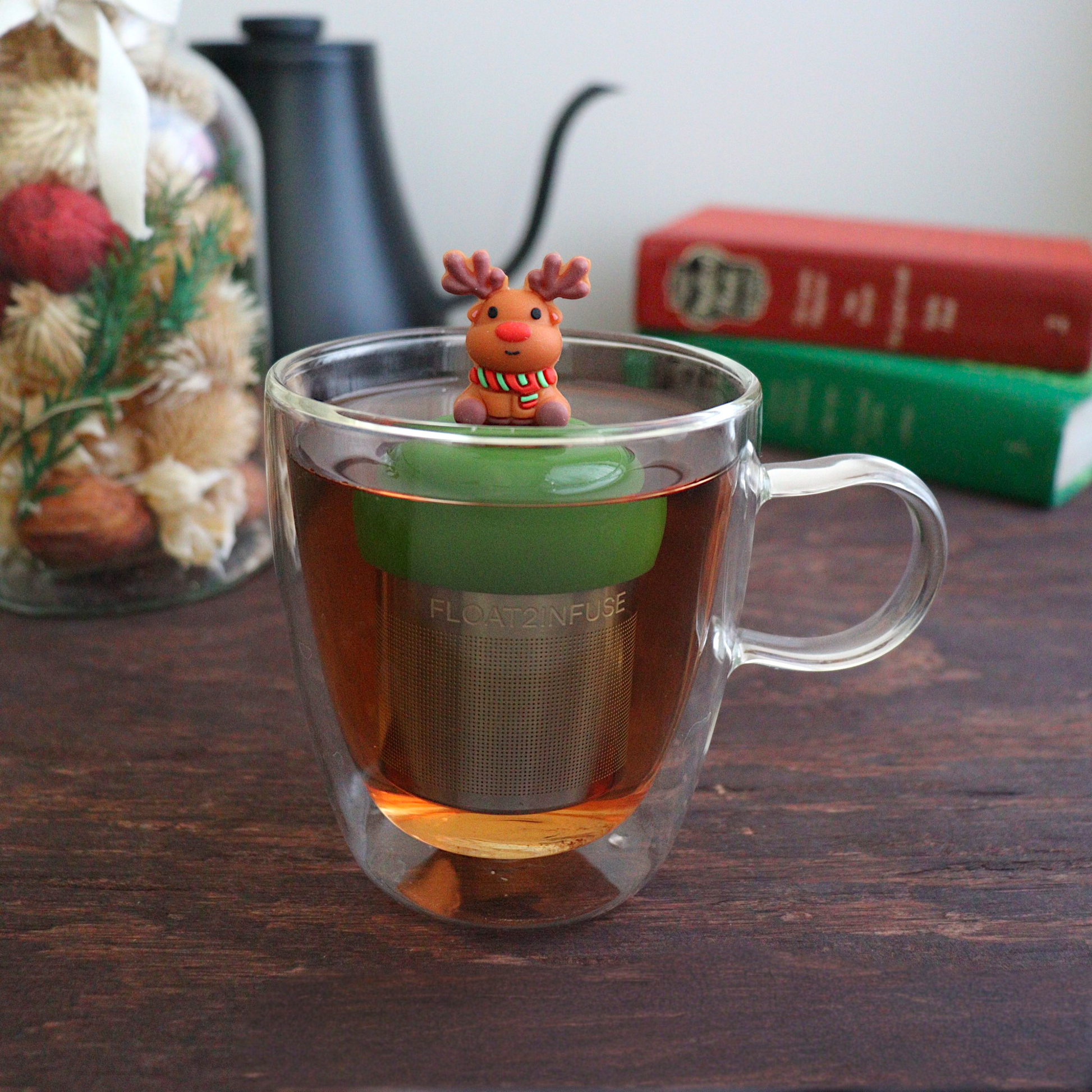 Reindeer with red nose wearing a green and red striped scarf tea infuser for loose leaf tea with a fine mesh stainless steel tea strainer 
that reads FLOAT2INFUSE on a dark green base floating in a glass tea mug with books, a tea kettle,  and Christmas decorations in the background