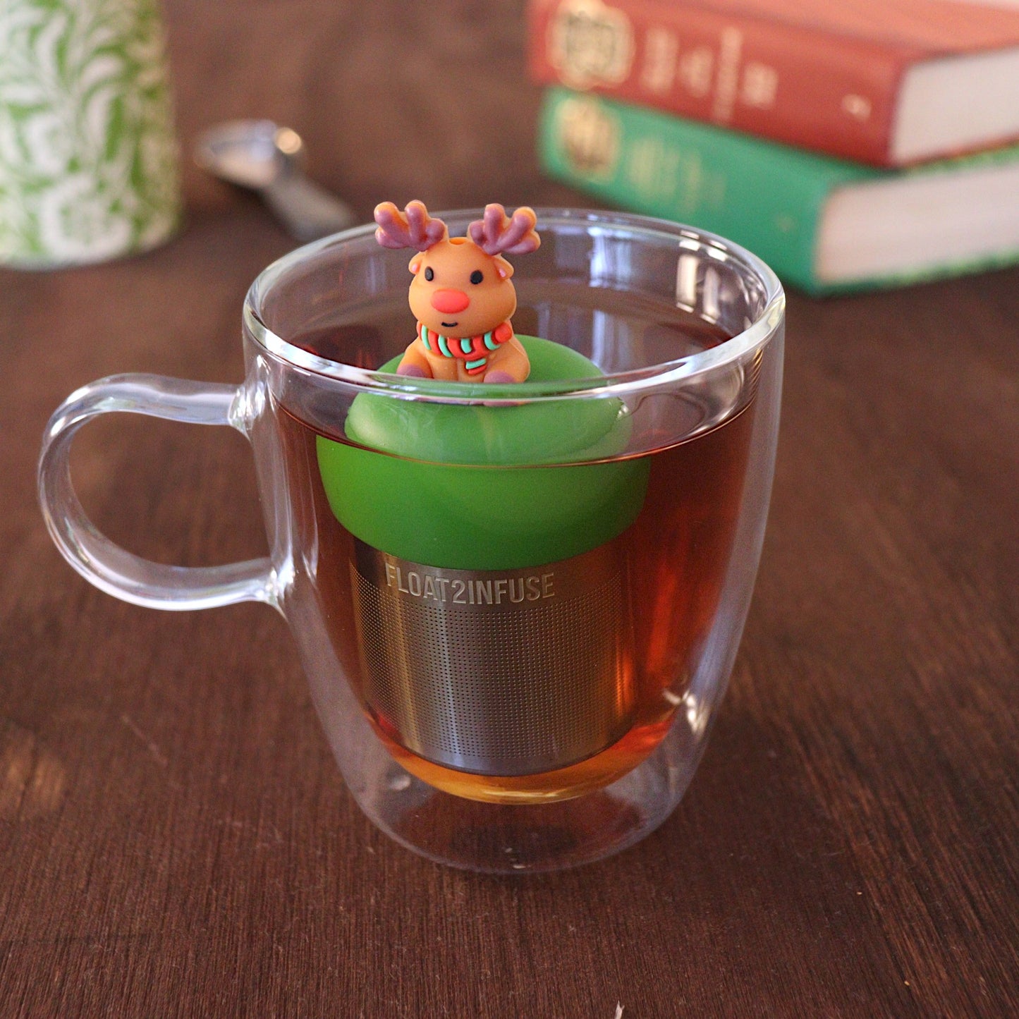 Reindeer with red nose wearing a green and red striped scarf tea infuser for loose leaf tea with a fine mesh stainless steel tea strainer 
that reads FLOAT2INFUSE on a dark green base floating in a glass tea mug with a tea canister and books in the background