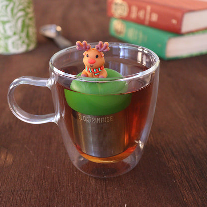 Reindeer with red nose wearing a green and red striped scarf tea infuser for loose leaf tea with a fine mesh stainless steel tea strainer 
that reads FLOAT2INFUSE on a dark green base floating in a glass tea mug with a tea canister and books in the background