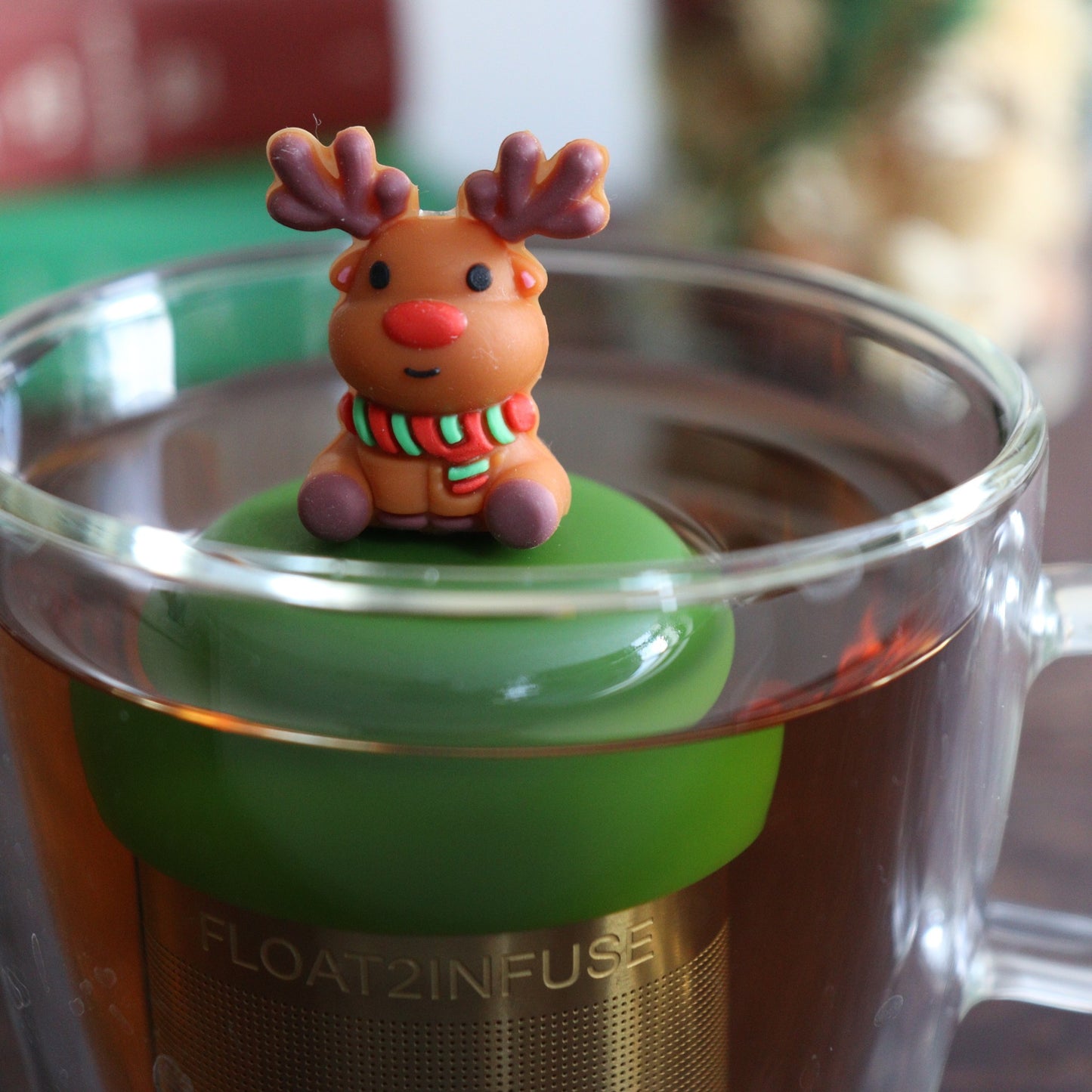 Reindeer with red nose wearing a green and red striped scarf tea infuser for loose leaf tea with a fine mesh stainless steel tea strainer 
that reads FLOAT2INFUSE on a dark green base floating in a glass tea mug with books and Christmas decorations in the background