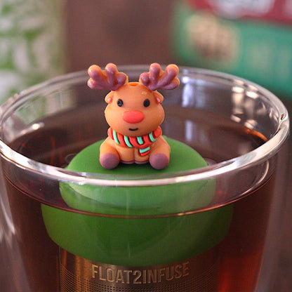 Reindeer with red nose wearing a green and red striped scarf tea infuser for loose leaf tea with a fine mesh stainless steel tea strainer 
that reads FLOAT2INFUSE on a dark green base floating in a glass tea mug with a tea canister and books in the background