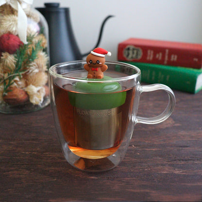 happy gingerbread man with a red bowtie and wearing a red and white santa hat  tea infuser for loose leaf tea with a fine mesh stainless steel tea strainer that reads FLOAT2INFUSE on a dark green base with a tea canister and books in the background
