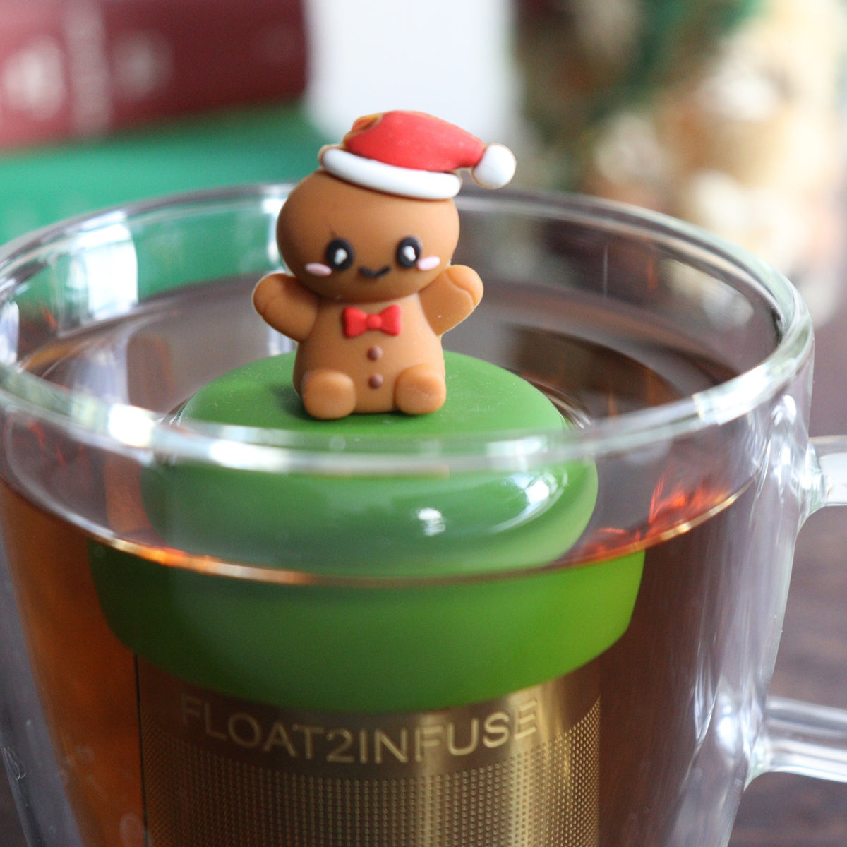 happy gingerbread man with a red bowtie and wearing a red and white santa hat infuser for loose leaf tea with a fine mesh stainless steel tea strainer that reads FLOAT2INFUSE on a dark green base with a tea canister and books in the background