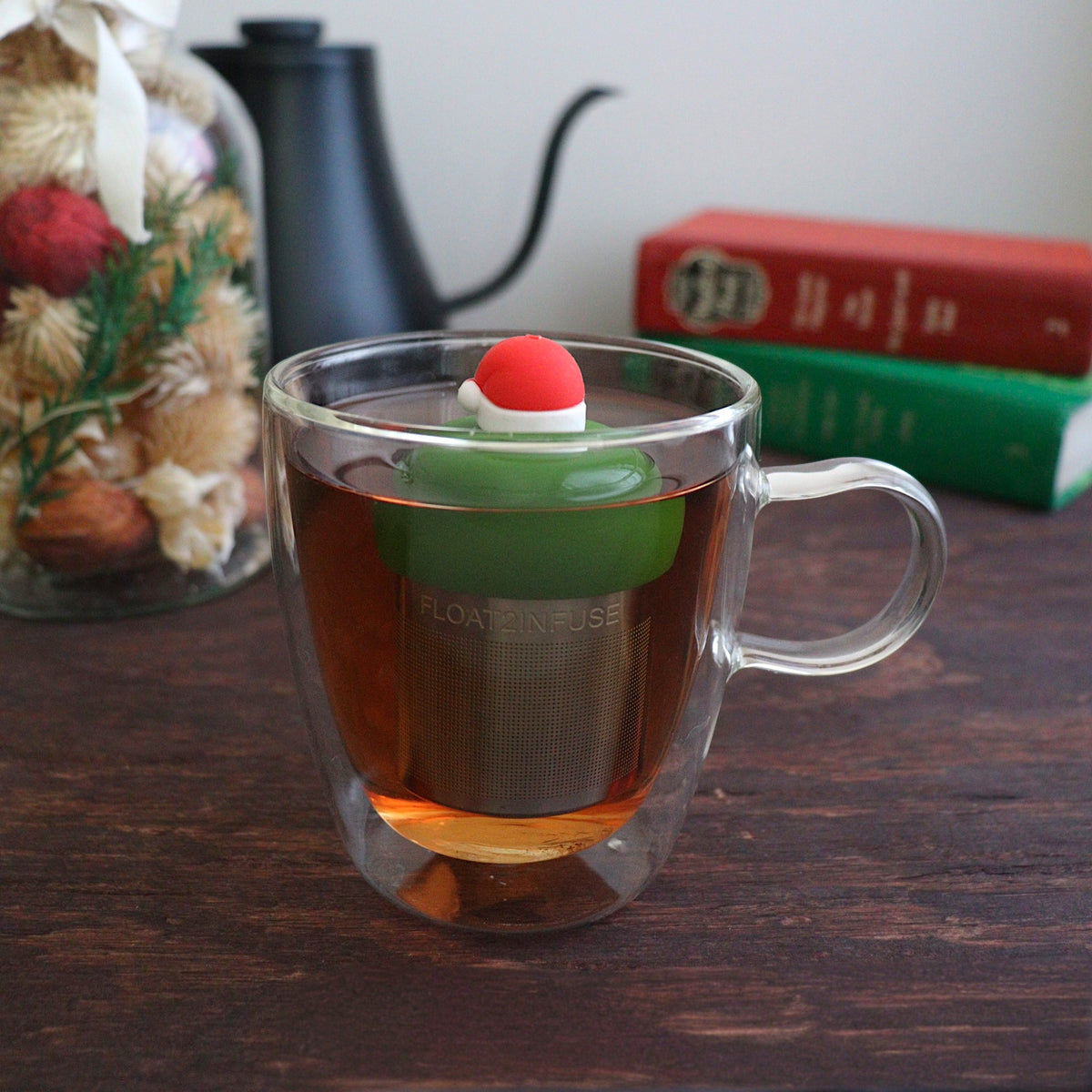 red santa hat with white trim tea infuser for loose leaf tea with a fine mesh stainless steel tea strainer 
that reads FLOAT2INFUSE on a dark green base floating in a glass tea mug with books and Christmas decorations in the background