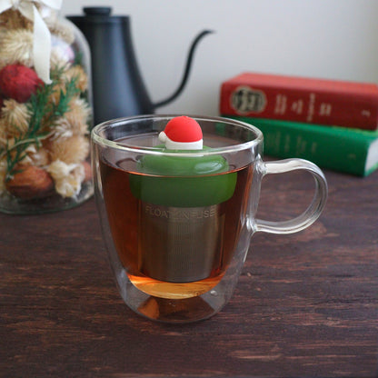 red santa hat with white trim tea infuser for loose leaf tea with a fine mesh stainless steel tea strainer 
that reads FLOAT2INFUSE on a dark green base floating in a glass tea mug with books and Christmas decorations in the background