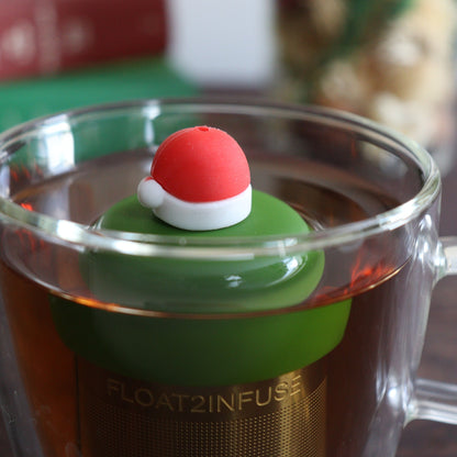 red santa hat with white trim tea infuser for loose leaf tea with a fine mesh stainless steel tea strainer 
that reads FLOAT2INFUSE on a dark green base floating in a glass tea mug with books and Christmas decorations in the background
