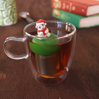 Cozy white polar bear wearing a red and white santa hat and a red scarf tea infuser for loose leaf tea with a fine mesh stainless steel tea strainer 
that reads FLOAT2INFUSE on a dark green base with books and a tea canister in the background