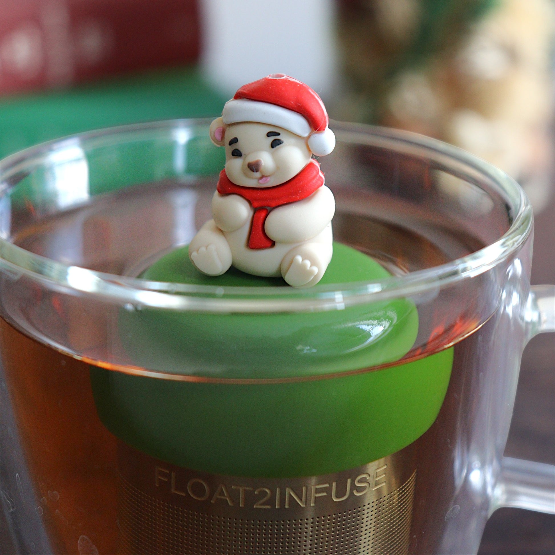 Cozy white polar bear wearing a red and white santa hat and a red scarf tea infuser for loose leaf tea with a fine mesh stainless steel tea strainer 
that reads FLOAT2INFUSE on a dark green base with books, a tea kettle, and christmas decorations in the background