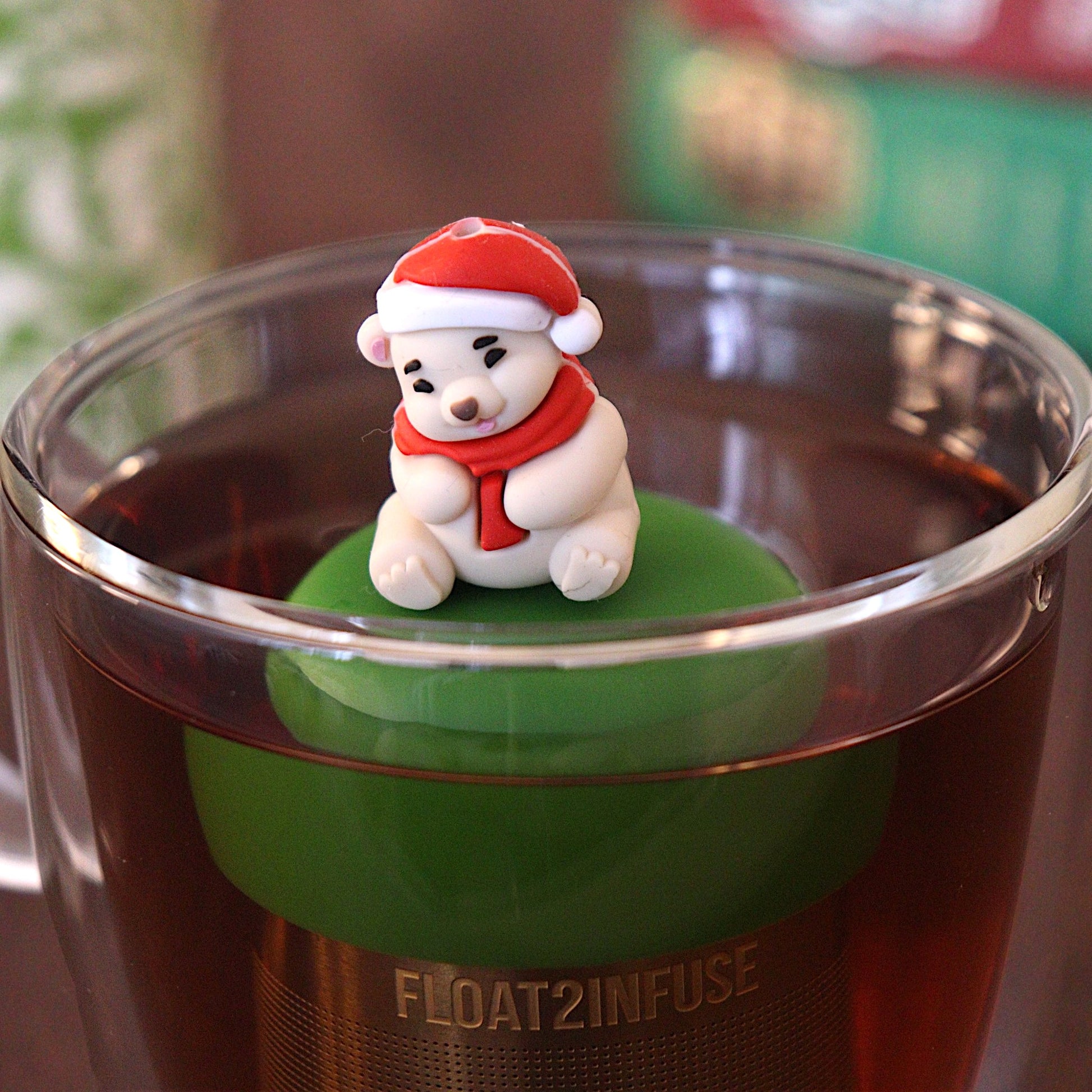 Cozy white polar bear wearing a red and white santa hat and a red scarf tea infuser for loose leaf tea with a fine mesh stainless steel tea strainer 
that reads FLOAT2INFUSE on a dark green base with books and a tea canister in the background