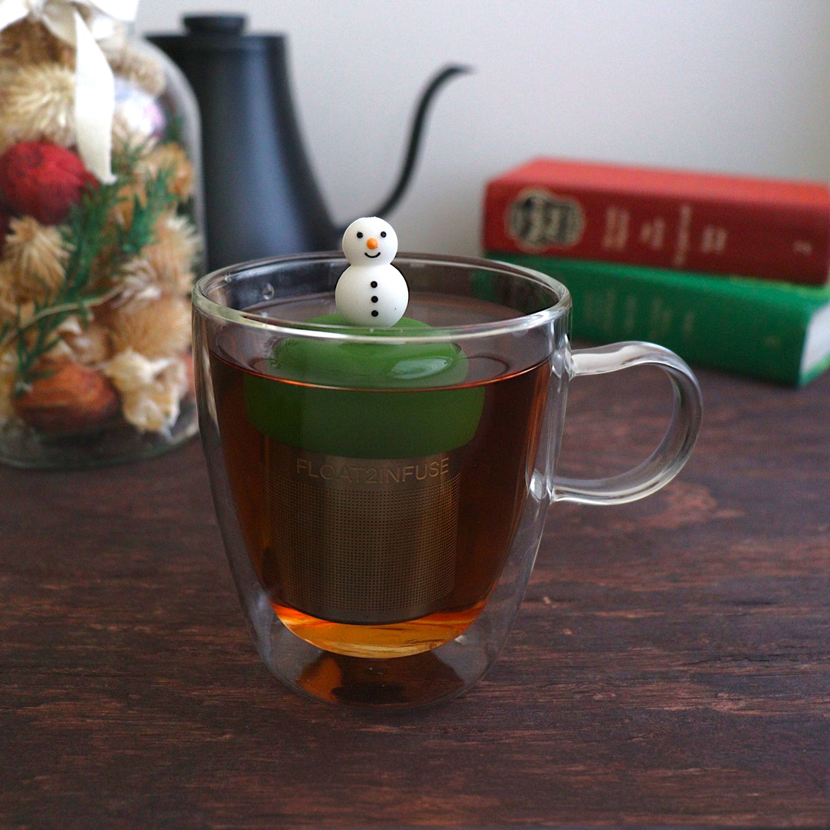 cute smiling snowman tea infuser for loose leaf tea with a fine mesh stainless steel tea strainer that reads FLOAT2INFUSE on a dark green base with books, a tea kettle, and christmas decorations in the background