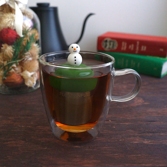 cute smiling snowman tea infuser for loose leaf tea with a fine mesh stainless steel tea strainer that reads FLOAT2INFUSE on a dark green base with books, a tea kettle, and christmas decorations in the background