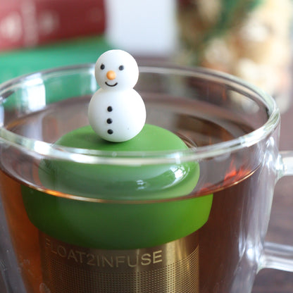 cute smiling snowman tea infuser for loose leaf tea with a fine mesh stainless steel tea strainer that reads FLOAT2INFUSE on a dark green base with books, a tea kettle, and christmas decorations in the background