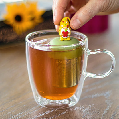 gnome with long white beard wearing yellow with red, white and green polka dots on the hat and holding a red apple animal tea infuser for loose leaf tea with a fine mesh stainless steel tea strainer that reads FLOAT2INFUSE on a green base floating in a glass tea mug with flowers in the background
