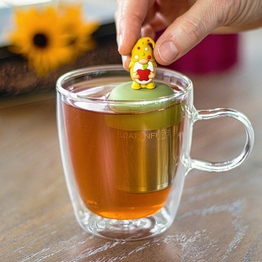gnome with long white beard wearing yellow with red, white and green polka dots on the hat and holding a red apple animal tea infuser for loose leaf tea with a fine mesh stainless steel tea strainer that reads FLOAT2INFUSE on a green base floating in a glass tea mug with flowers in the background