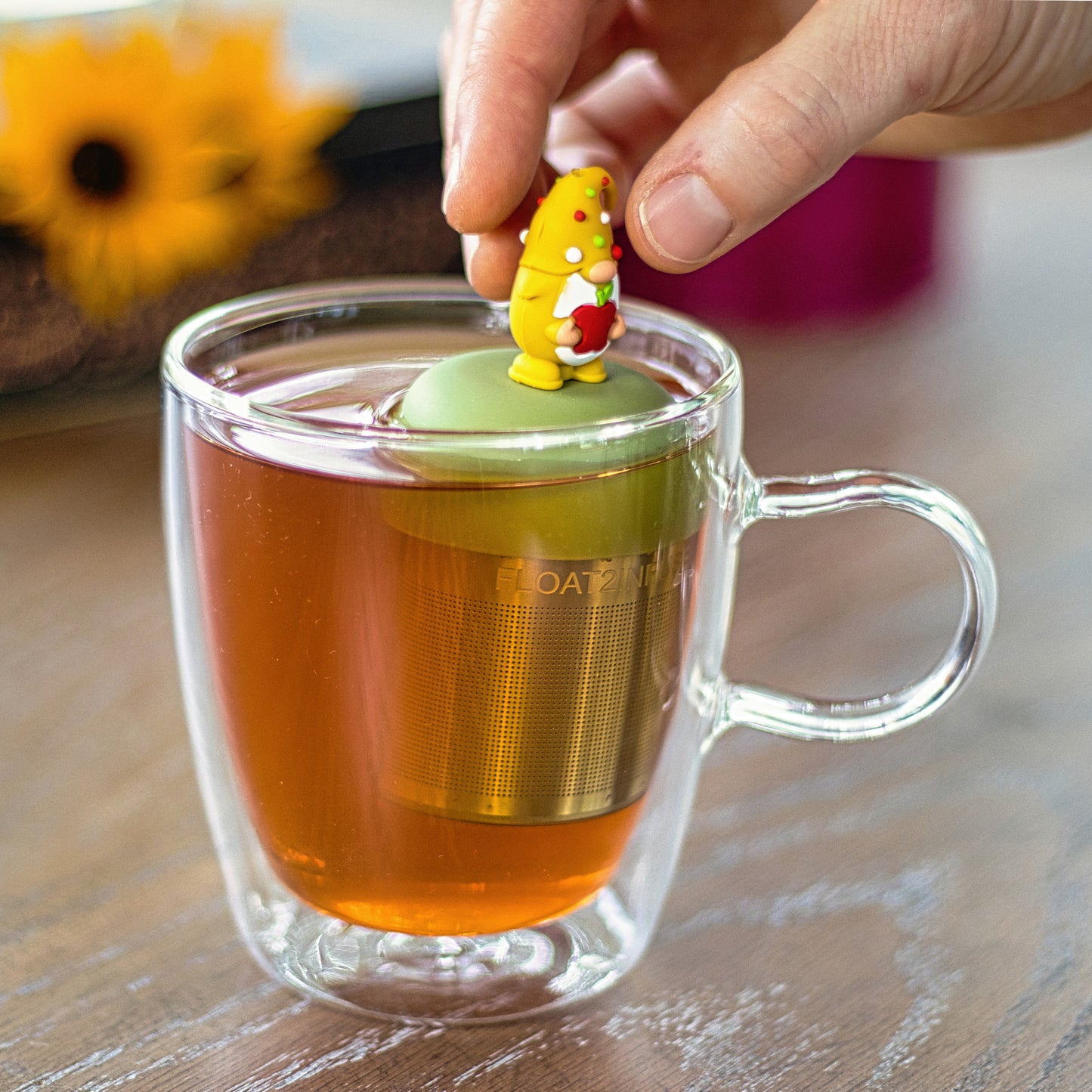 gnome with long white beard wearing yellow with red, white and green polka dots on the hat and holding a red apple animal tea infuser for loose leaf tea with a fine mesh stainless steel tea strainer that reads FLOAT2INFUSE on a green base floating in a glass tea mug with flowers in the background