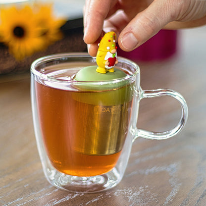 gnome with long white beard wearing yellow with red, white and green polka dots on the hat and holding a red apple animal tea infuser for loose leaf tea with a fine mesh stainless steel tea strainer that reads FLOAT2INFUSE on a green base floating in a glass tea mug with flowers in the background