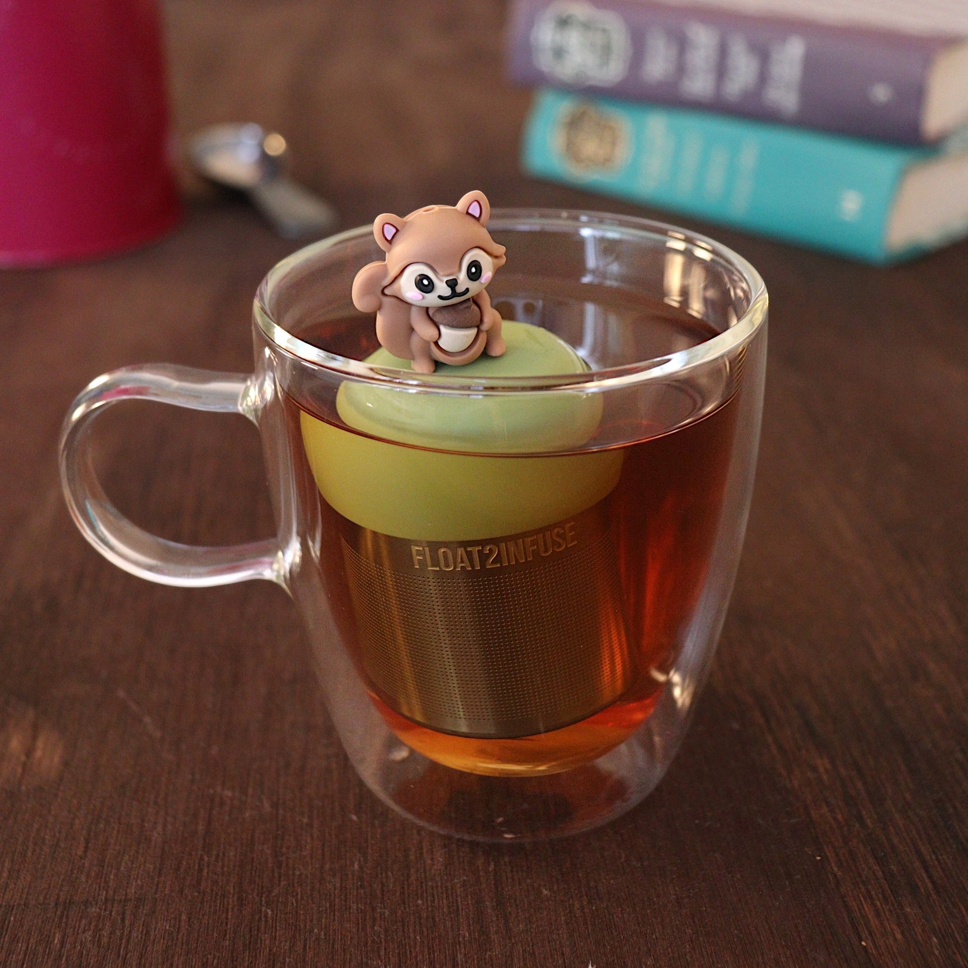 Cute 3D brown squirrel holding an acorn tea infuser for loose leaf tea with a fine mesh stainless steel tea strainer that reads FLOAT2INFUSE on a green base with a tea canister and books in the background
