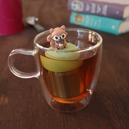 Cute 3D brown squirrel holding an acorn tea infuser for loose leaf tea with a fine mesh stainless steel tea strainer that reads FLOAT2INFUSE on a green base with a tea canister and books in the background