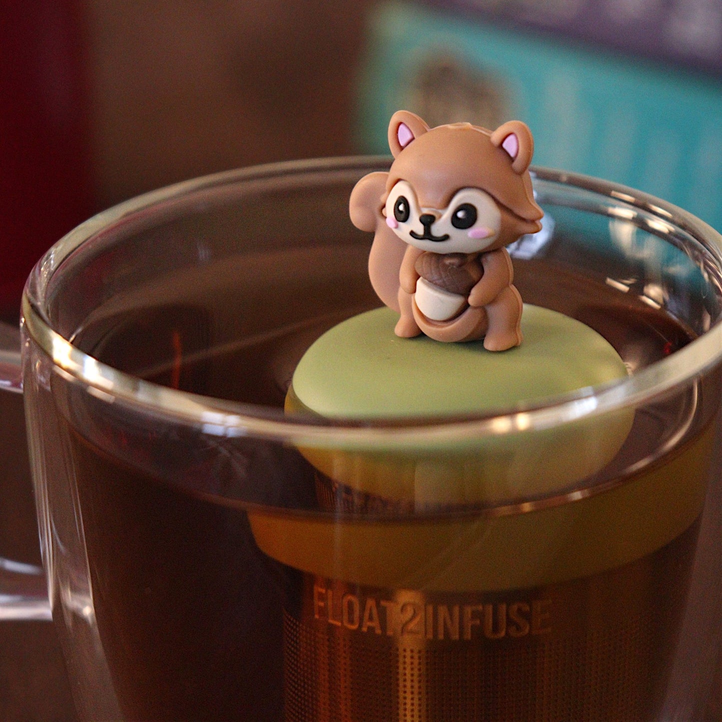 Cute 3D brown squirrel holding an acorn tea infuser for loose leaf tea with a fine mesh stainless steel tea strainer that reads FLOAT2INFUSE on a green base with a tea canister and books in the background