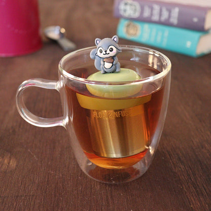 Cute 3d grey squirrel holding an acorn tea infuser for loose leaf tea with a fine mesh stainless steel tea strainer that reads FLOAT2INFUSE on a green base with a tea canister and books in the background