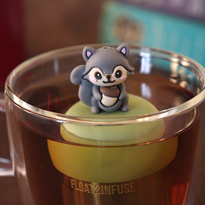 Cute 3d grey squirrel holding an acorn tea infuser for loose leaf tea with a fine mesh stainless steel tea strainer that reads FLOAT2INFUSE on a green base with a tea canister and books in the background