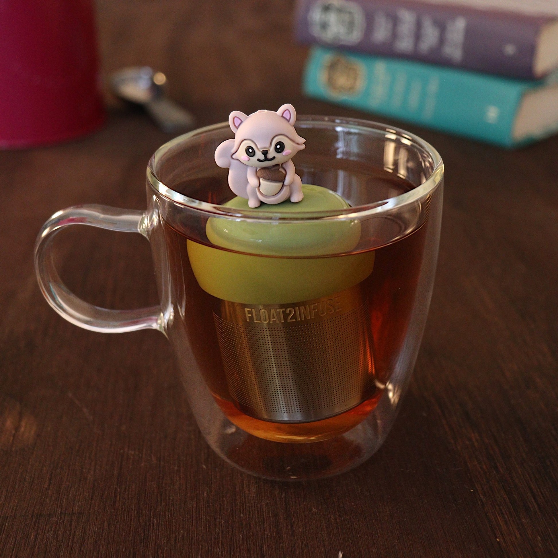 Cute 3D rose gold colored squirrel holding an acorn tea infuser for loose leaf tea with a fine mesh stainless steel tea strainer that reads FLOAT2INFUSE on a green base with a tea canister and books in the background