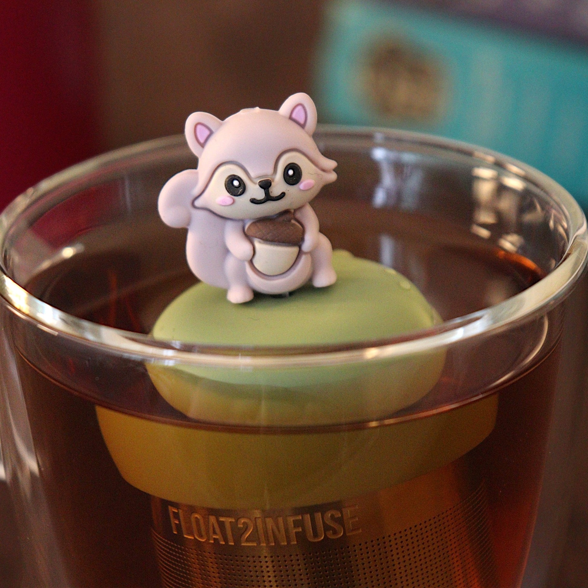 Cute 3D rose gold colored squirrel holding an acorn tea infuser for loose leaf tea with a fine mesh stainless steel tea strainer that reads FLOAT2INFUSE on a green base with a tea canister and books in the background