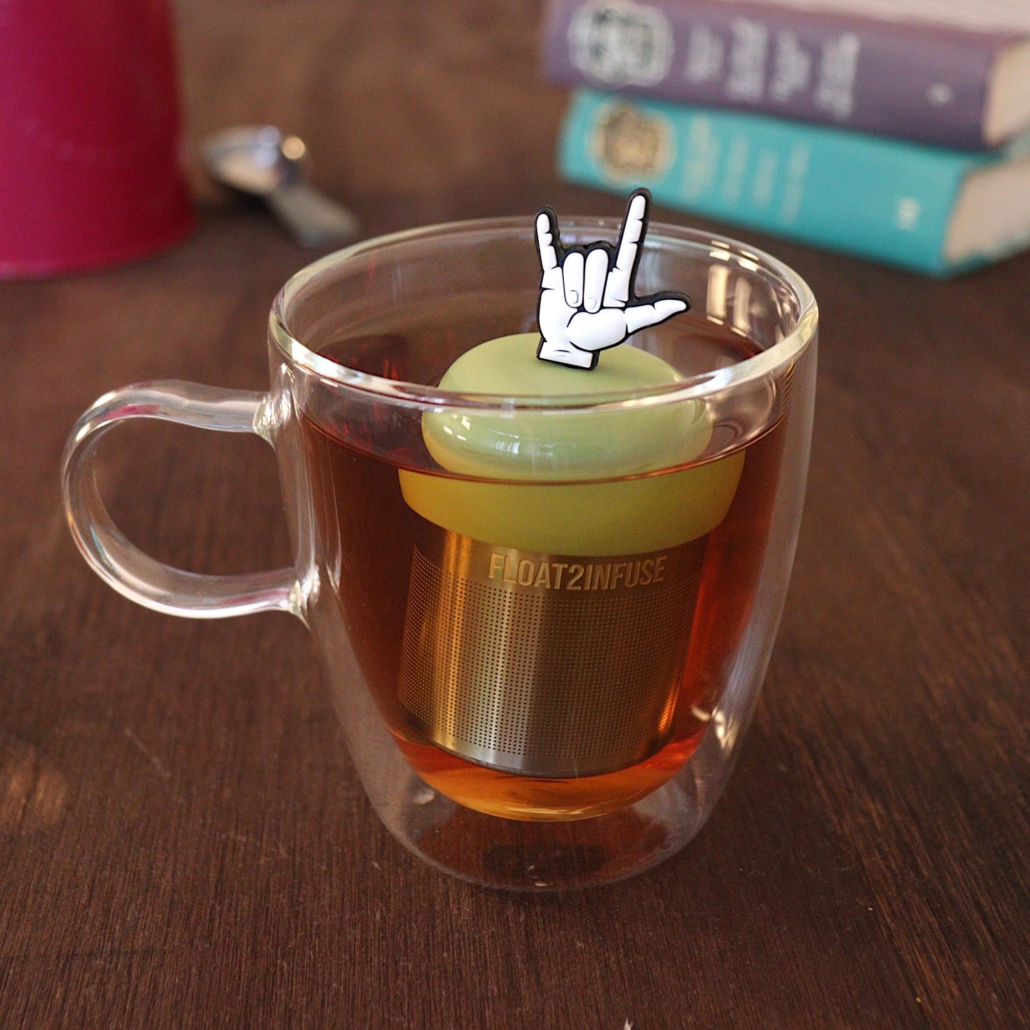 American Sign Language sign for "I Love You" tea infuser for loose leaf tea with a fine mesh stainless steel tea strainer that reads FLOAT2INFUSE on a green base with a tea canister and books in the background