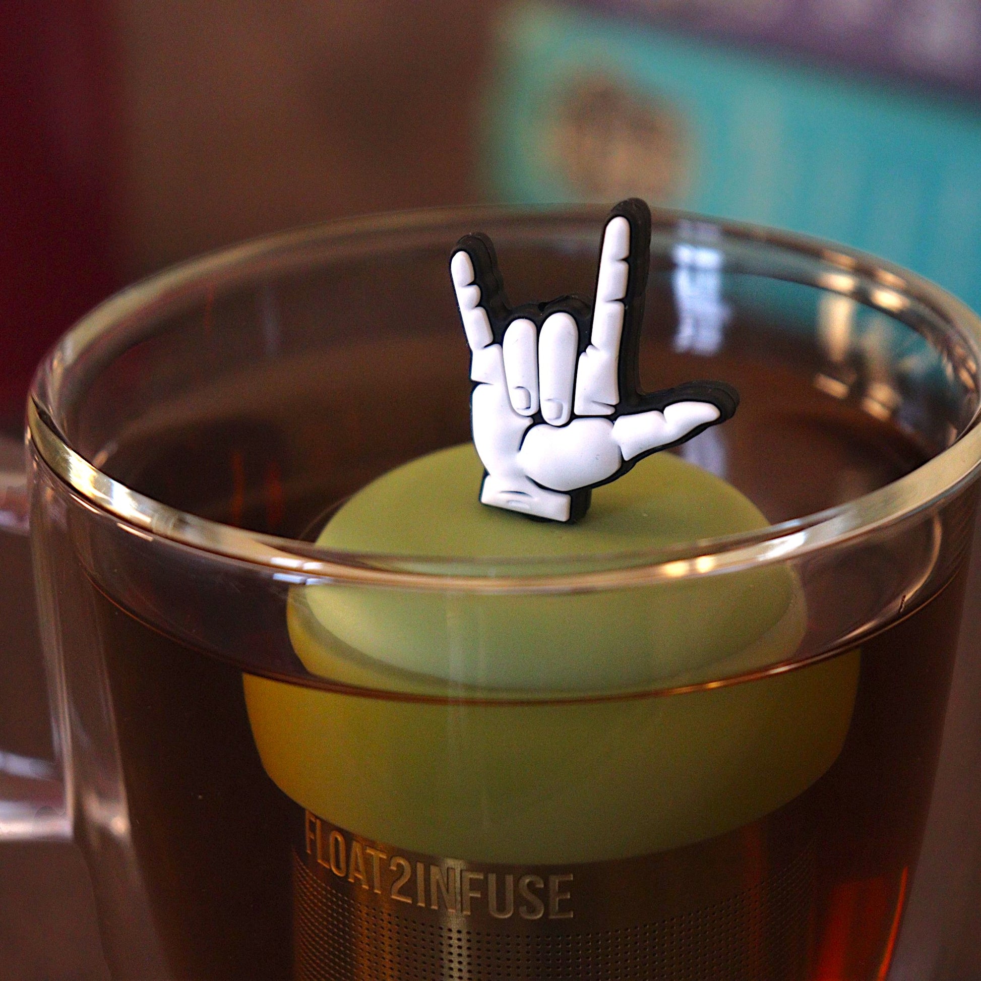 American Sign Language sign for "I Love You" tea infuser for loose leaf tea with a fine mesh stainless steel tea strainer that reads FLOAT2INFUSE on a green base with a tea canister and books in the background