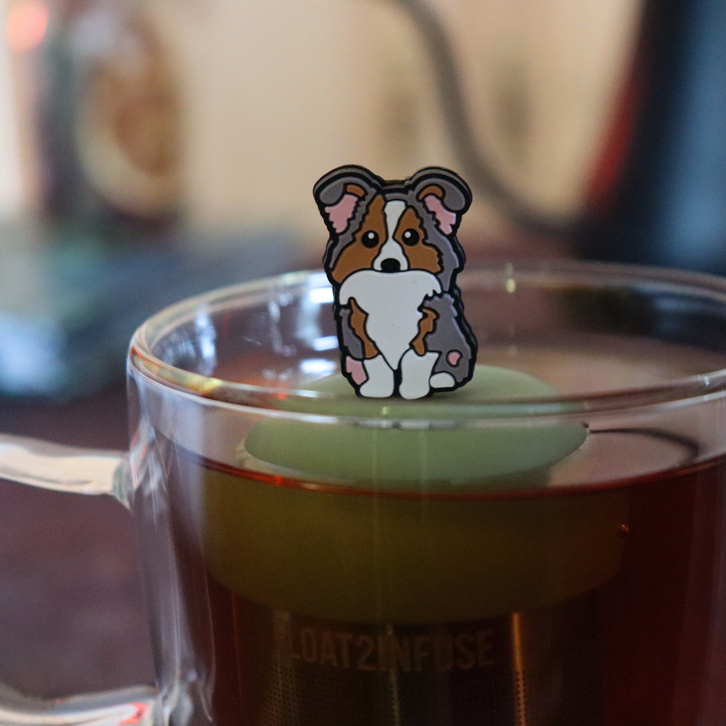 grey and brown and white Australian shepherd dog animal tea infuser for loose leaf tea with a fine mesh stainless steel tea strainer that reads FLOAT2INFUSE on a green base floating in a glass tea mug with tea kettle in the background