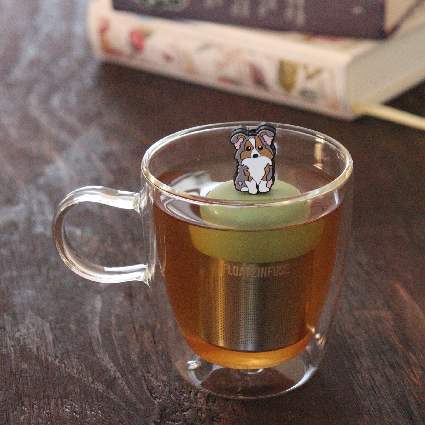 grey and brown and white Australian shepherd dog animal tea infuser for loose leaf tea  with a fine mesh stainless steel tea infuser that reads FLOAT2INFUSE on a green base floating in a glass tea mug with books in the background