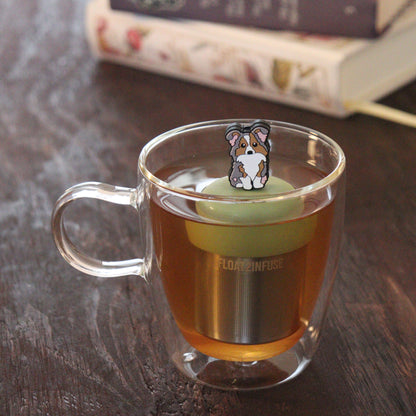 grey and brown and white Australian shepherd dog animal tea infuser for loose leaf tea  with a fine mesh stainless steel tea infuser that reads FLOAT2INFUSE on a green base floating in a glass tea mug with books in the background