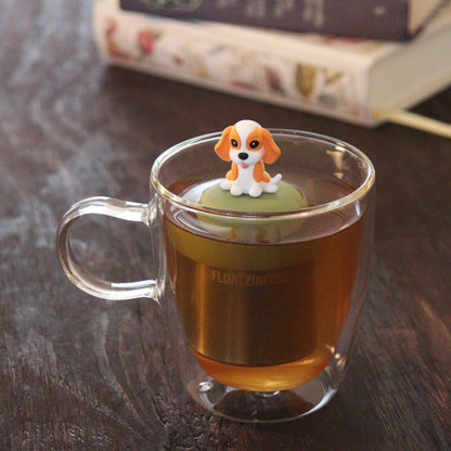 orange and white beagle animal tea infuser for loose leaf tea  with a fine mesh stainless steel tea infuser that reads FLOAT2INFUSE on a green base floating in a glass tea mug with books in the background