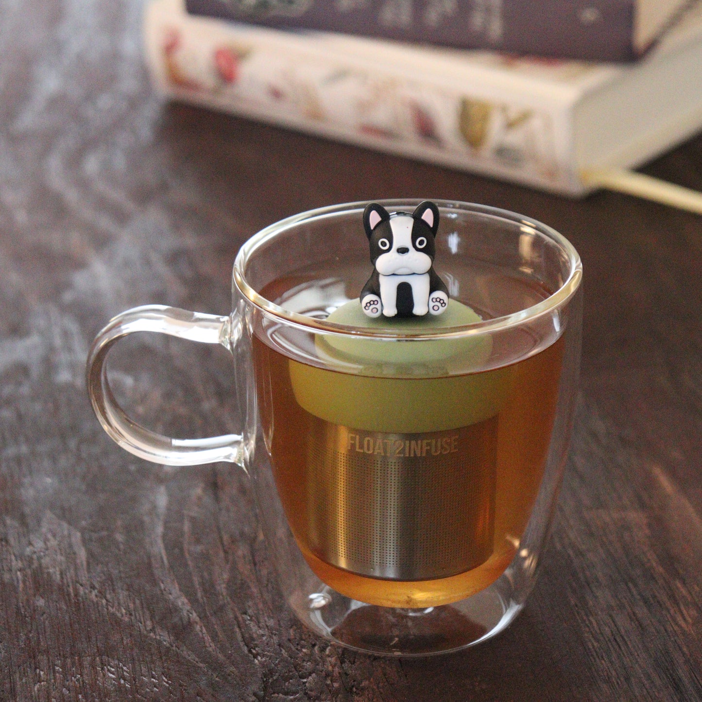 black and white terrier dog animal tea infuser for loose leaf tea on a green base with a fine mesh stainless steel tea infuser that reads FLOAT2INFUSE floating in a glass tea mug with books in the background