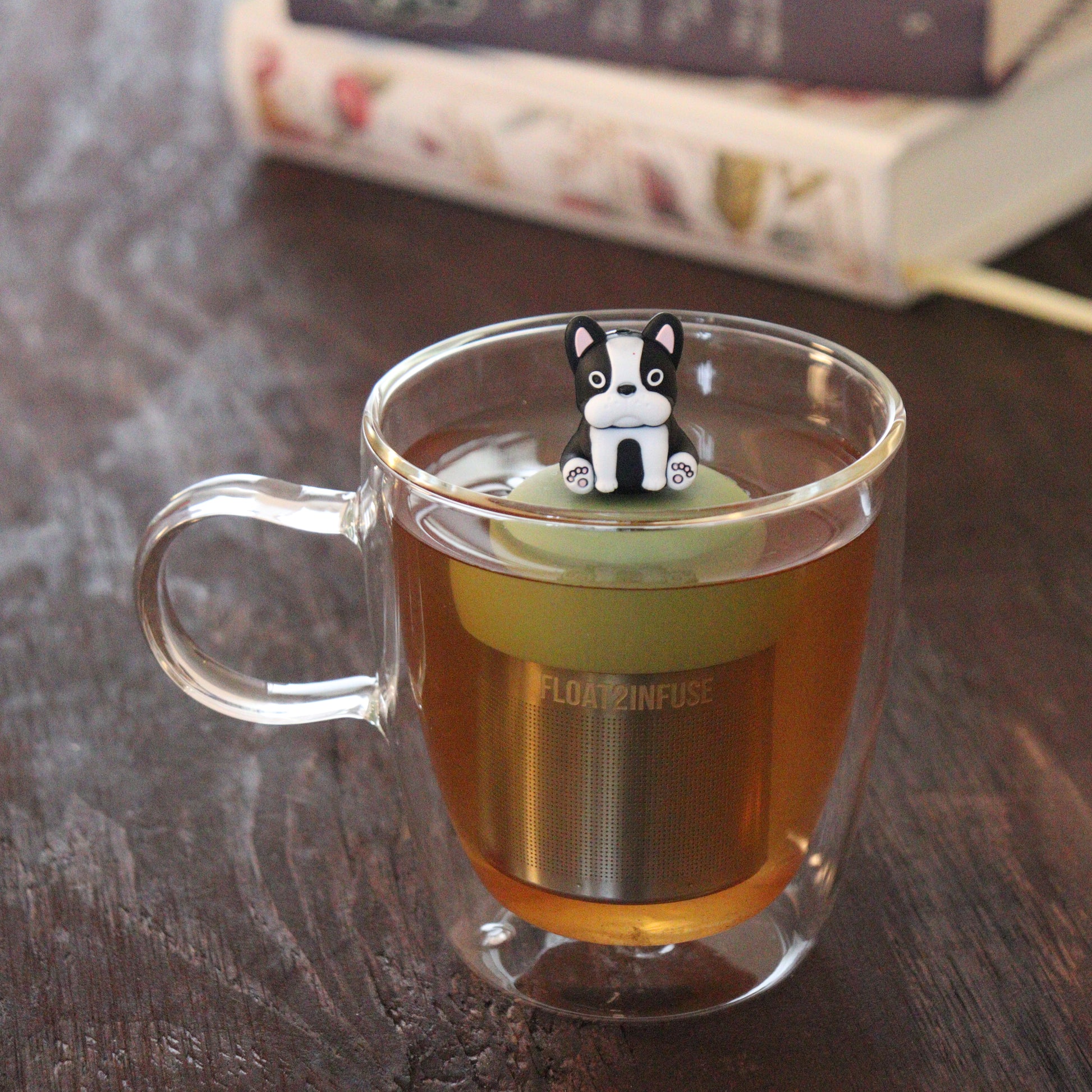 black and white terrier dog animal tea infuser for loose leaf tea on a green base with a fine mesh stainless steel tea infuser that reads FLOAT2INFUSE floating in a glass tea mug with books in the background