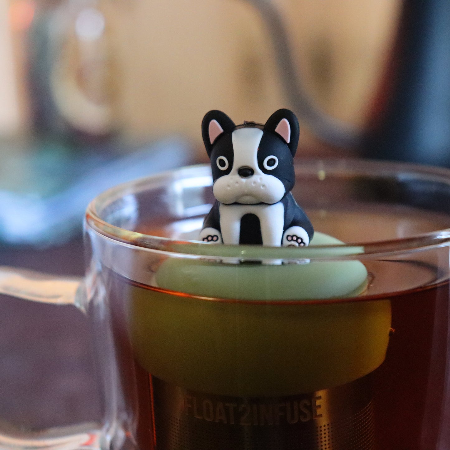 black and white terrier dog animal tea infuser for loose leaf tea on a green base with a fine mesh stainless steel tea strainer that reads FLOAT2INFUSE floating in a glass tea mug with tea kettle in the background