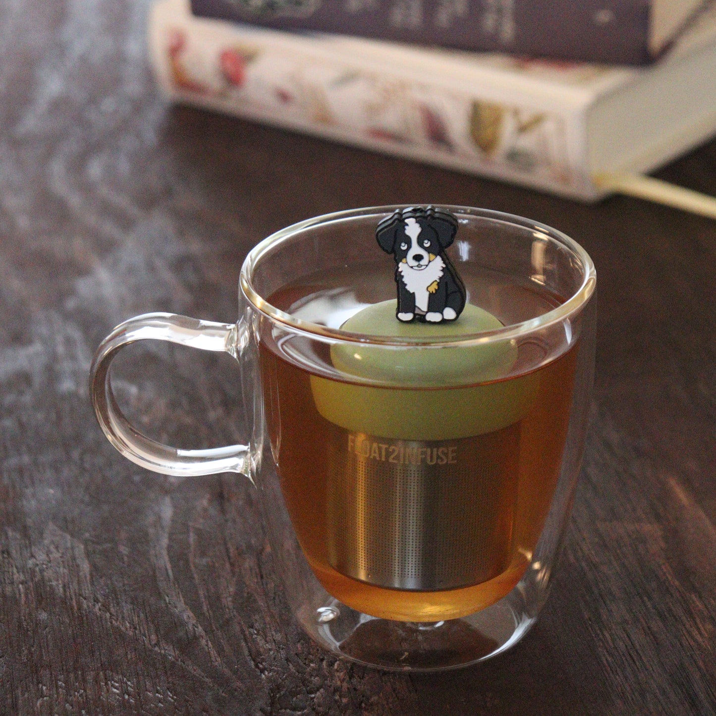 black and white border collie dog animal tea infuser for loose leaf tea  with a fine mesh stainless steel tea infuser that reads FLOAT2INFUSE on a green base floating in a glass tea mug with books in the background