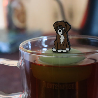 brown boxer dog animal tea infuser for loose leaf tea on a green base with a fine mesh stainless steel tea strainer that reads FLOAT2INFUSE floating in a glass tea mug with tea kettle in the background
