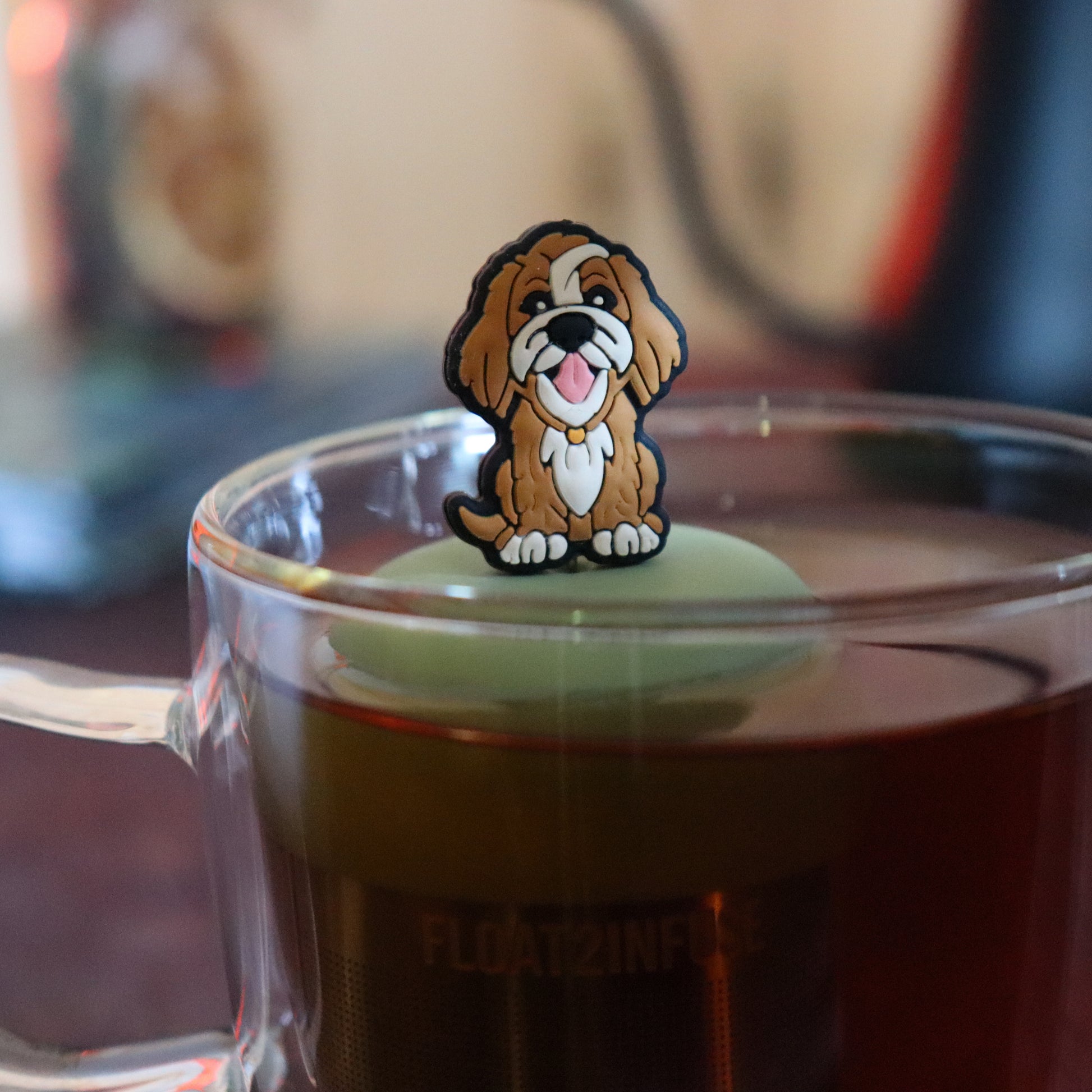 brown golden retriever dog animal tea infuser for loose leaf tea on with a fine mesh stainless steel tea strainer that reads FLOAT2INFUSE a green base floating in a glass tea mug with tea kettle in the background
