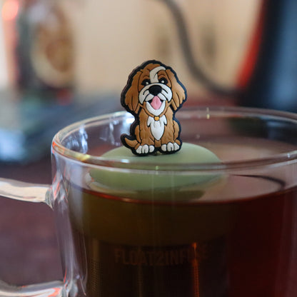 brown golden retriever dog animal tea infuser for loose leaf tea on with a fine mesh stainless steel tea strainer that reads FLOAT2INFUSE a green base floating in a glass tea mug with tea kettle in the background