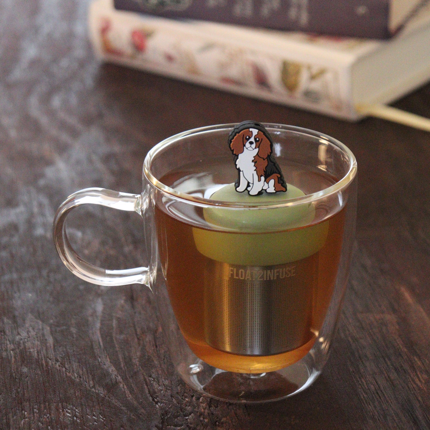 brown and white cocker spaniel dog animal tea infuser for loose leaf tea on a green base with a fine mesh stainless steel tea infuser that reads FLOAT2INFUSE floating in a glass tea mug with books in the background