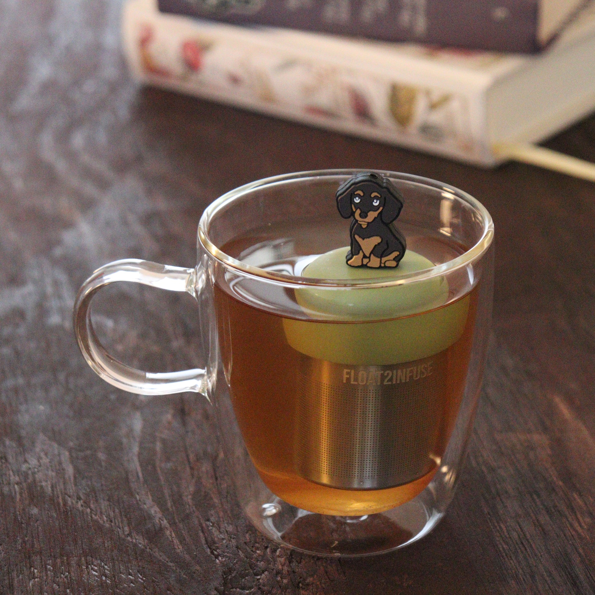 black and brown dachshund dog animal tea infuser for loose leaf tea  with a fine mesh stainless steel tea infuser that reads FLOAT2INFUSE on a green base floating in a glass tea mug with books in the background