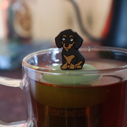 black and brown dachshund dog animal tea infuser for loose leaf tea with a fine mesh stainless steel tea strainer that reads FLOAT2INFUSE on a green base floating in a glass tea mug with tea kettle in the background