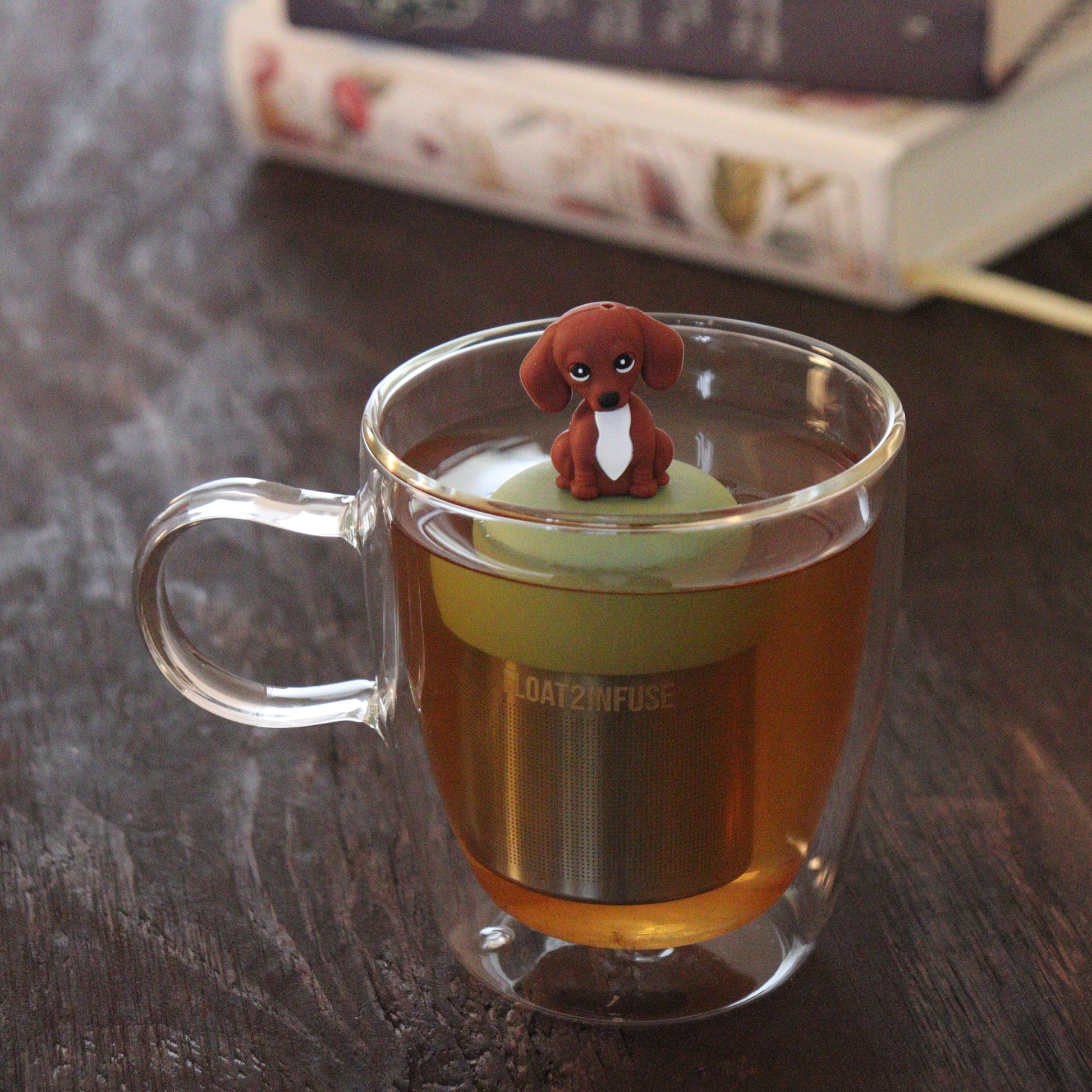 brown and white dachshund puppy dog animal tea infuser for loose leaf tea  with a fine mesh stainless steel tea infuser that reads FLOAT2INFUSE on a green base floating in a glass tea mug with books in the background