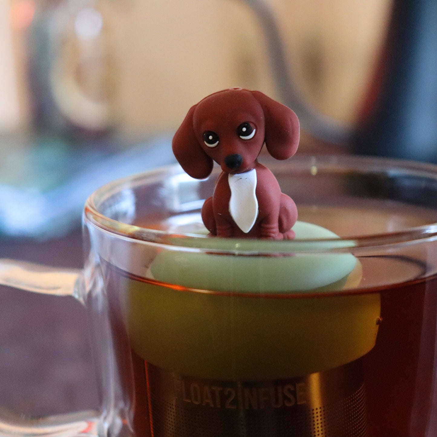 brown dachshund puppy dog animal tea infuser for loose leaf tea  with a fine mesh stainless steel tea strainer that reads FLOAT2INFUSE on a green base floating in a glass tea mug with tea kettle in the background