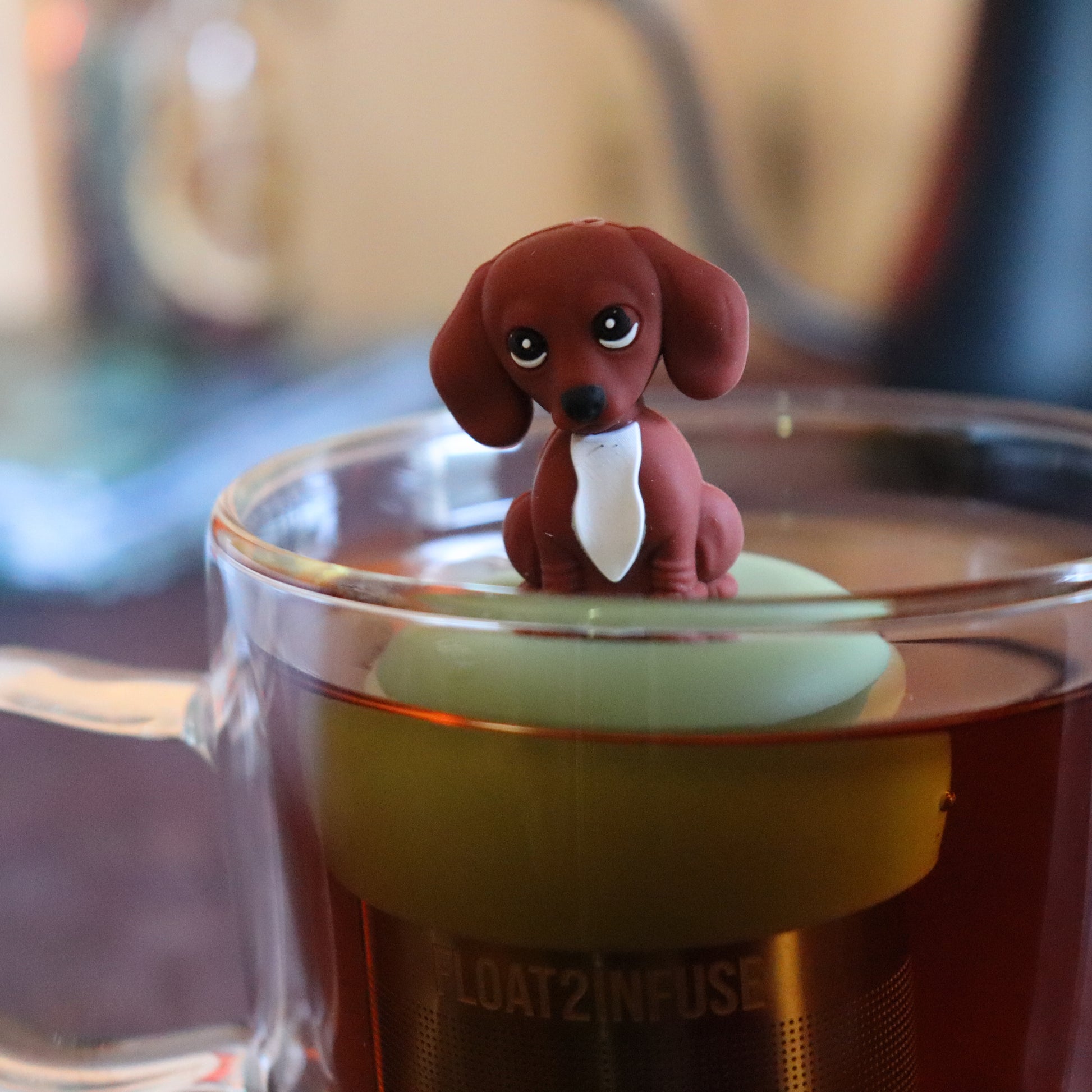 brown dachshund puppy dog animal tea infuser for loose leaf tea  with a fine mesh stainless steel tea strainer that reads FLOAT2INFUSE on a green base floating in a glass tea mug with tea kettle in the background