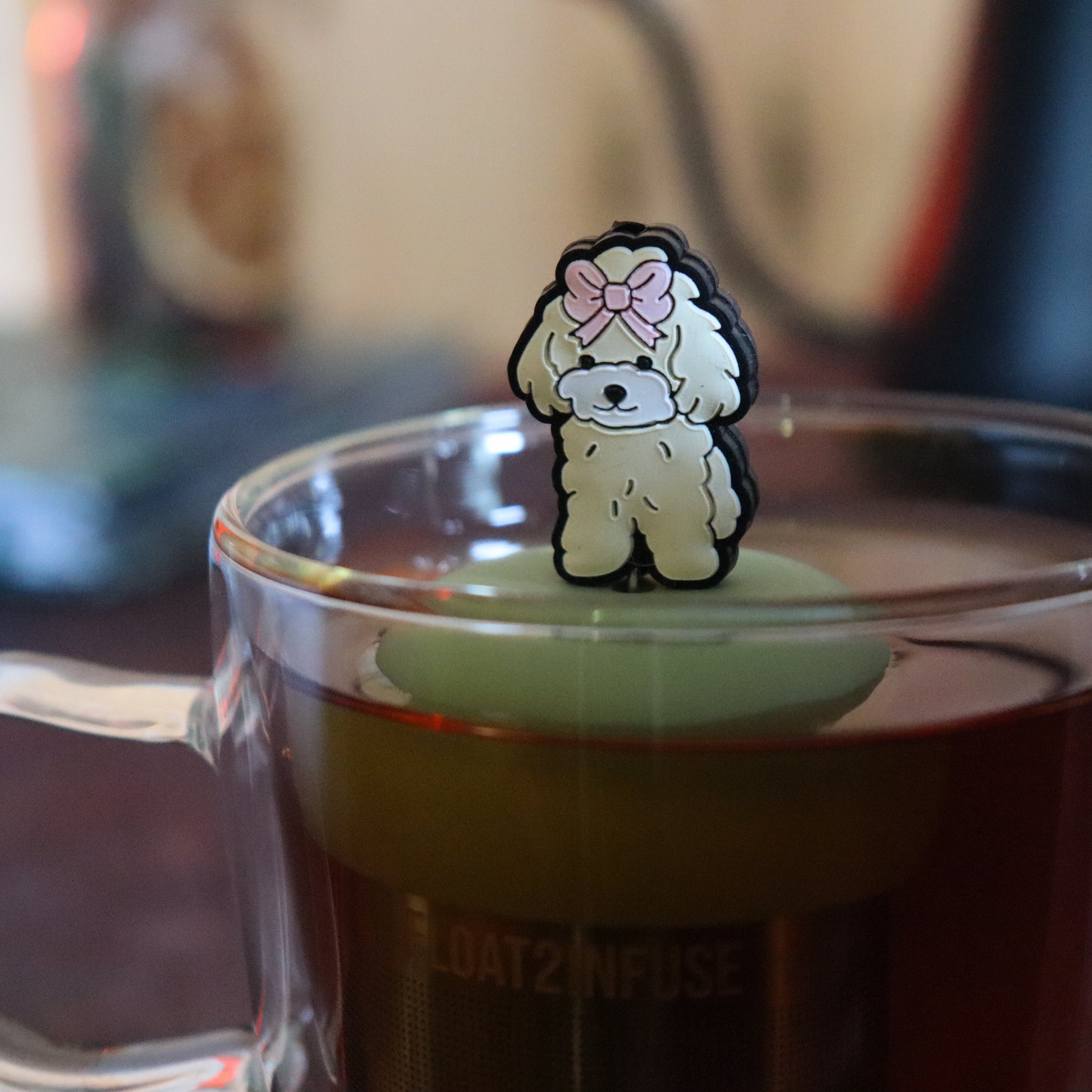 pink bow poodle dog animal tea infuser for loose leaf tea on a green base with a fine mesh stainless steel tea strainer that reads FLOAT2INFUSE floating in a glass tea mug with tea kettle in the background