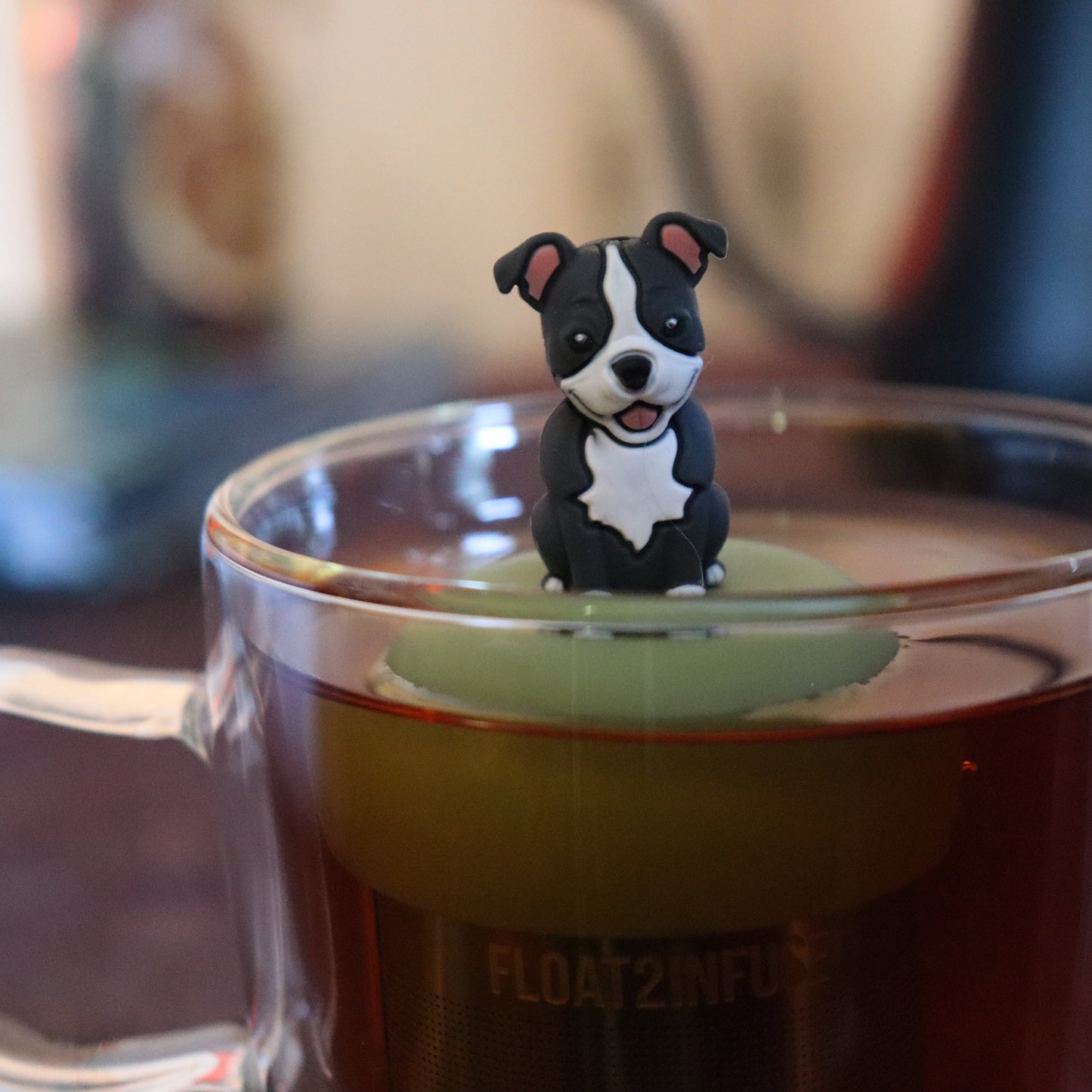 grey and white pit bull puppy dog animal tea infuser for loose leaf tea with a fine mesh stainless steel tea strainer that reads FLOAT2INFUSE on a green base floating in a glass tea mug with tea kettle in the background