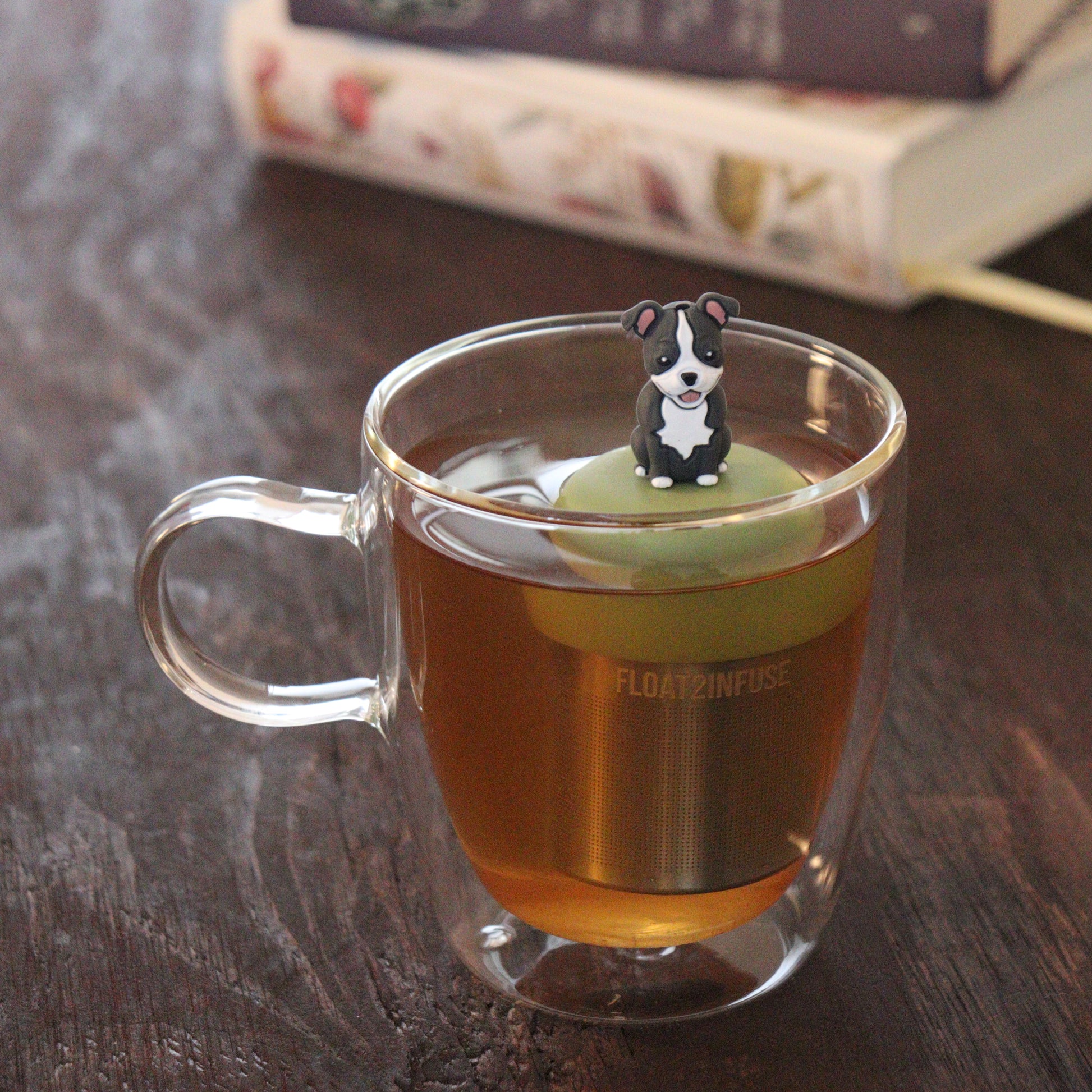 grey and white pit bull puppy dog animal tea infuser for loose leaf tea  with a fine mesh stainless steel tea infuser that reads FLOAT2INFUSE on a green base floating in a glass tea mug with books in the background